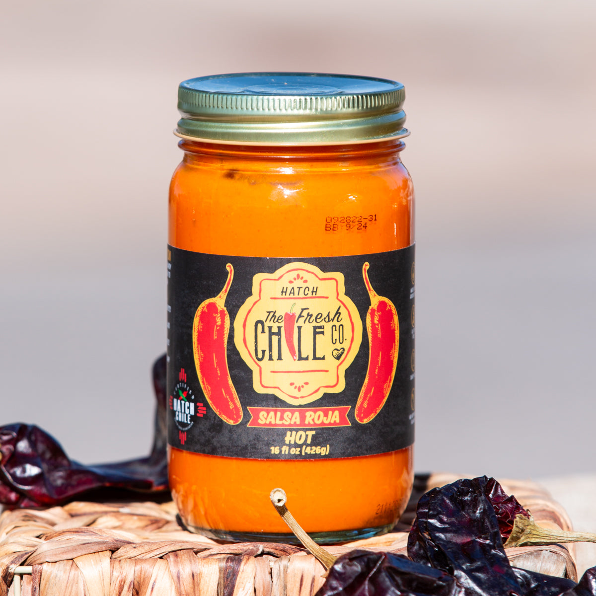 A jar of Hatch Jalapeño Salsa Roja from Hatch New Mexico, stands on a wicker mat surrounded by dried chile peppers. The label features bright red peppers and indicates