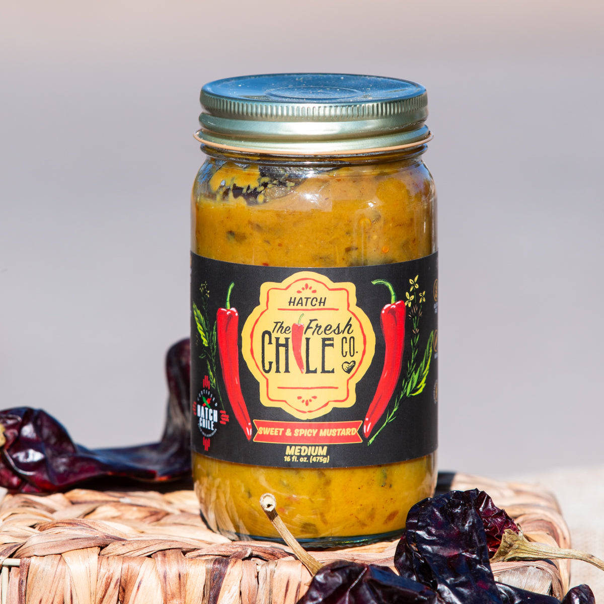 A jar of Sweet & Spicy Hatch Chile Mustard on a woven mat, flanked by dried chilies, with a neutral background.