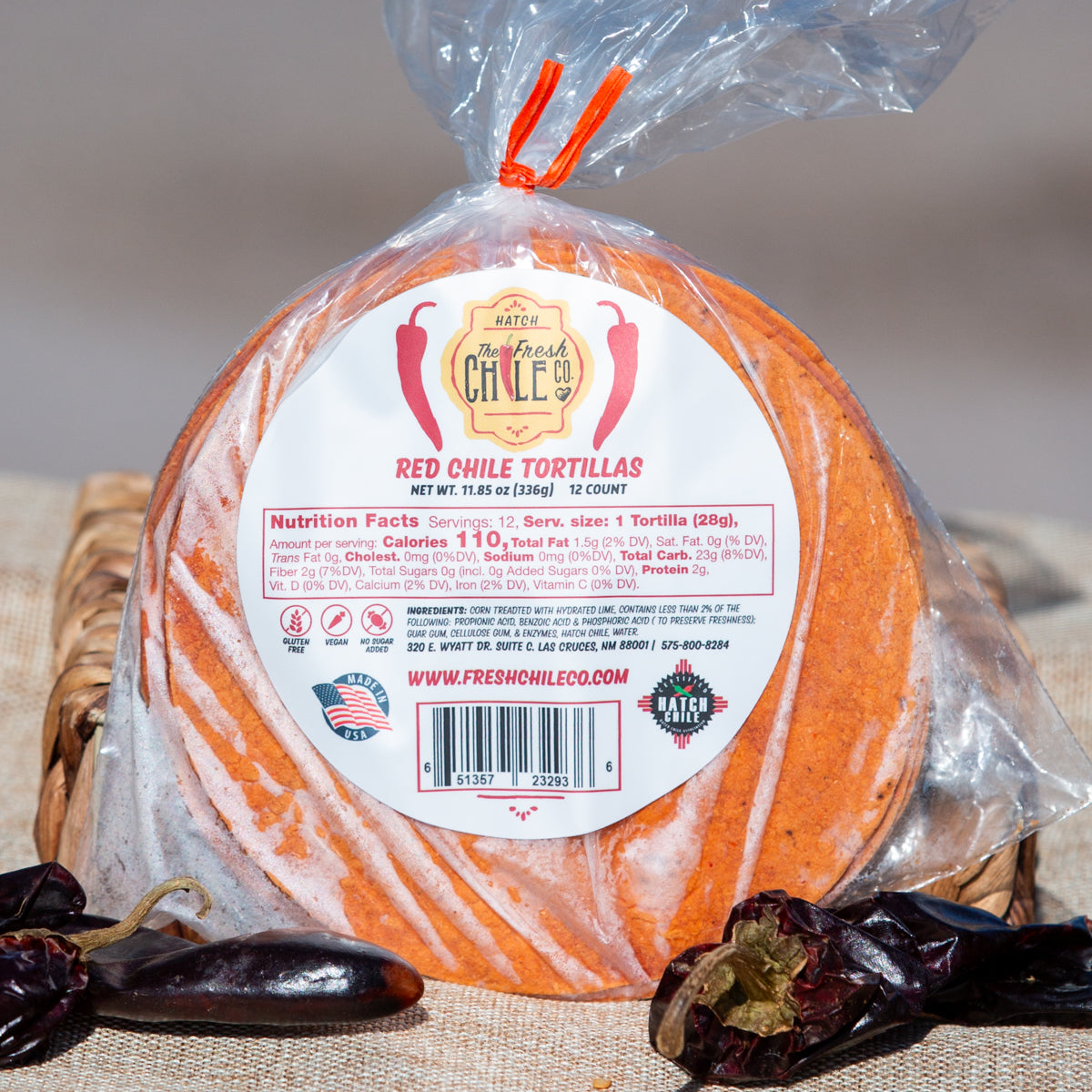 A pack of Red Chile Corn Tortillas in a clear plastic bag with a label, surrounded by dried chile peppers on a neutral textile surface. The label features nutritional info and branding details.
