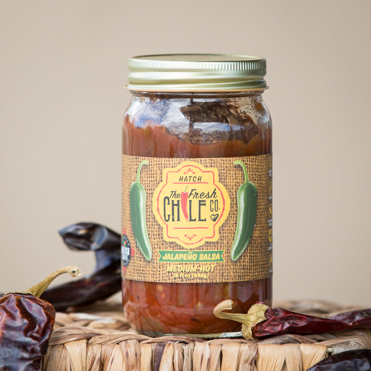 A jar of Hatch Jalapeño Salsa from The Fresh Chile Co. labeled "medium hot," recognized as a staple in New Mexican salsa, surrounded by dried chile peppers, on a woven basket against a neutral background.