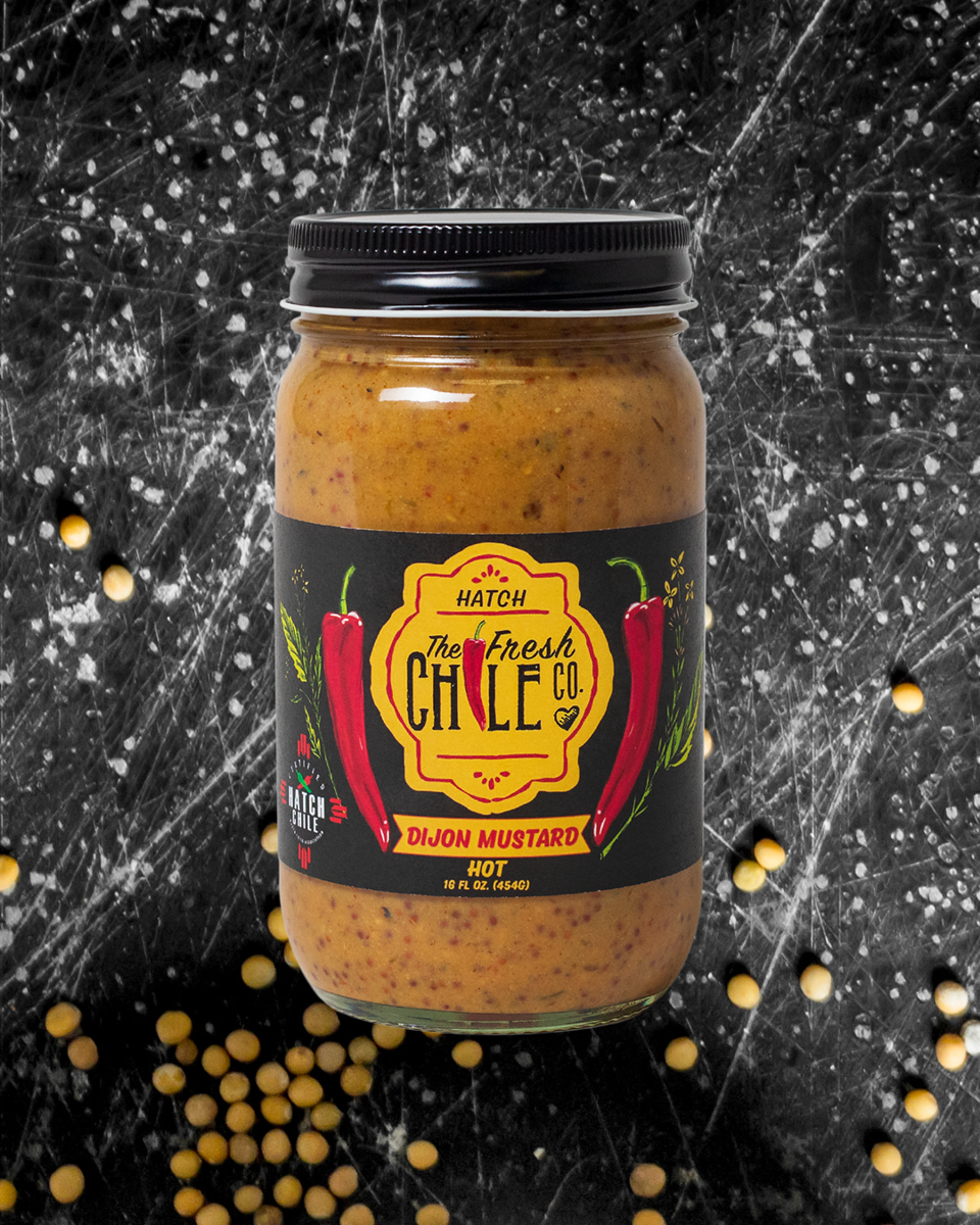 A jar of the Hatch Chile Dijon Mustard labeled "hot," featuring red chilies on the label, set against a dark, speckled background with scattered mustard seeds.