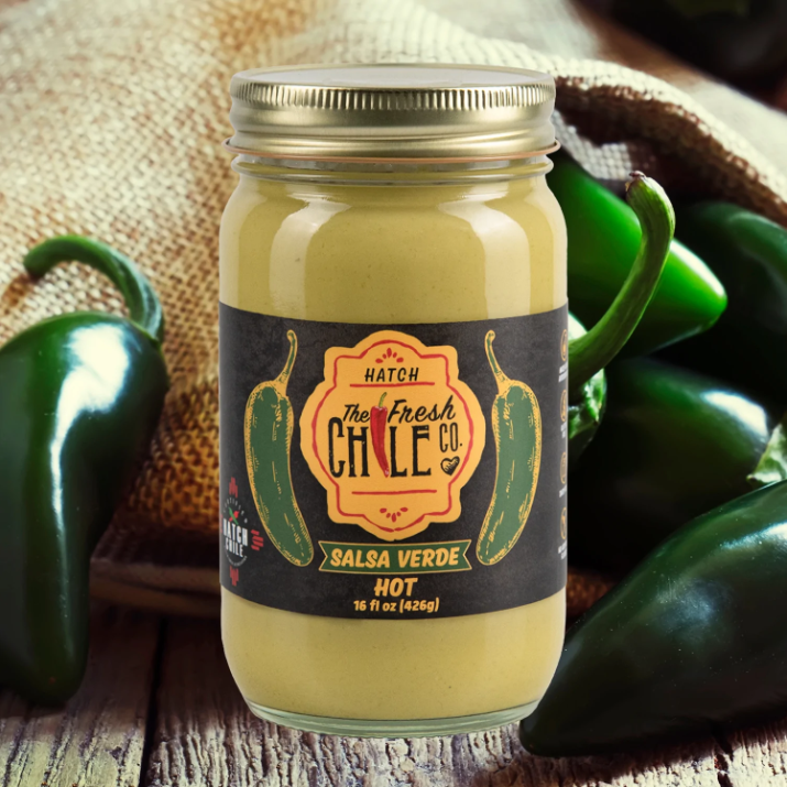 A jar of Hatch Jalapeño Salsa Verde from the fresh chile co., labeled "hot," placed on a wooden surface, surrounded by fresh green jalapeño peppers.