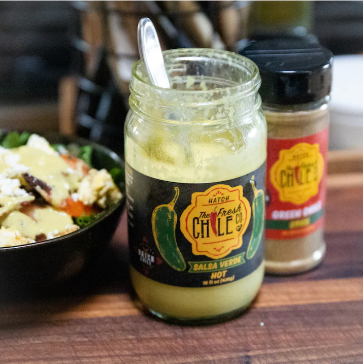 A glass jar of Hatch Jalapeño Salsa Verde on a wooden kitchen counter, with a spoon inside and a bottle of chili seasoning beside it. A salad bowl is visible in the background.