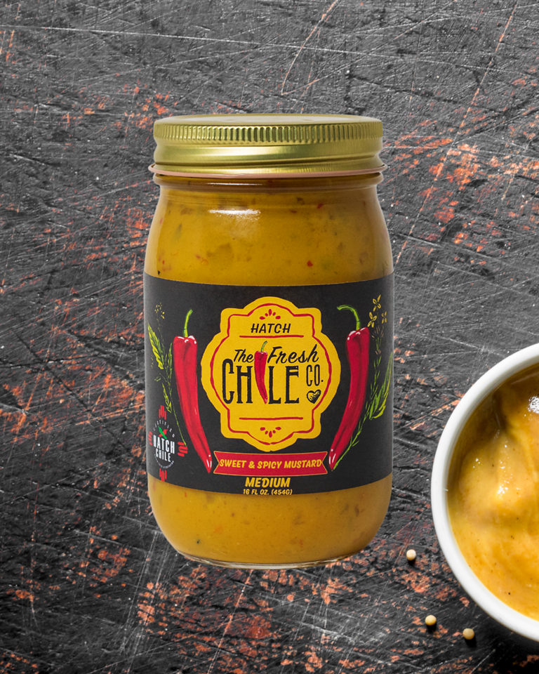 A jar of Sweet & Spicy Hatch Chile Mustard from The Fresh Chile Co. on a rough, dark textured background with a small bowl of the mustard visible to the side.