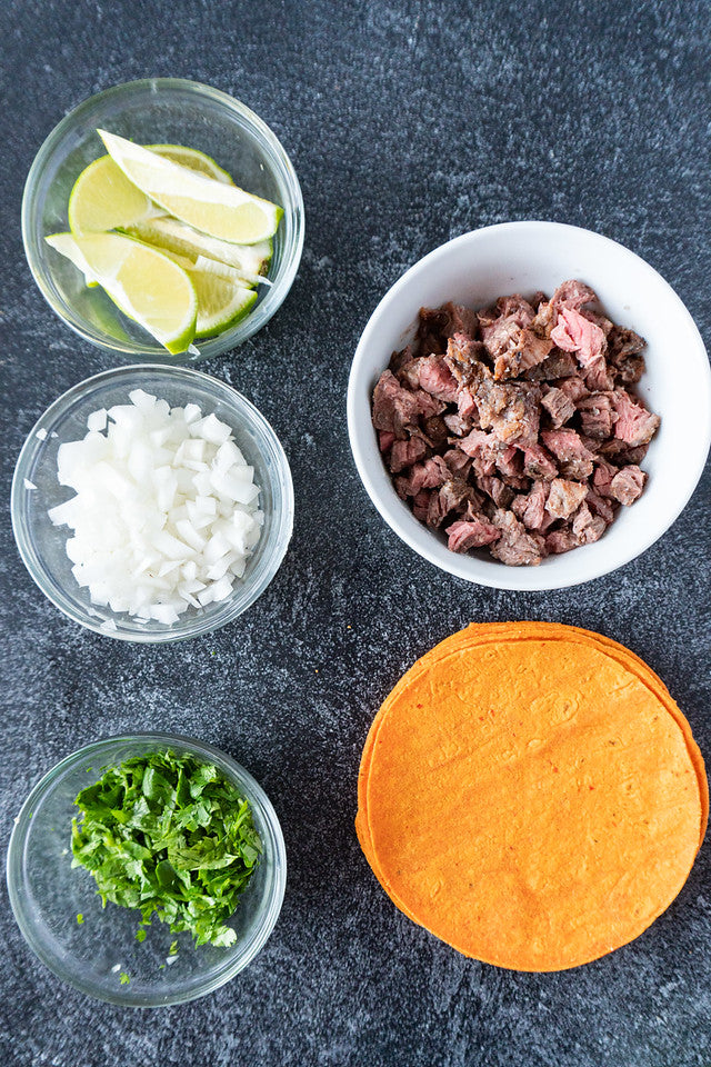 Overhead view of ingredients for tacos: diced beef, chopped onions, cilantro, lime wedges, and a stack of Red Chile Corn Tortillas, arranged neatly on a dark surface.