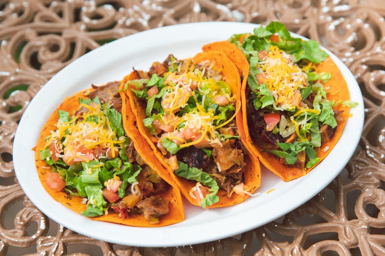 Three tacos with crispy Red Chile Corn Tortillas filled with seasoned meat, lettuce, tomatoes, and shredded cheese, served on a white plate over a decorative table.